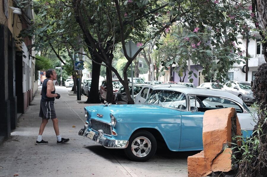 Classic blue car on busy street with greenery and casual walker.