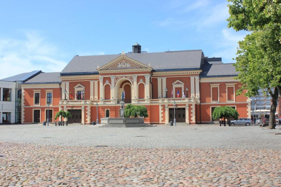 Grand historical building with columns, cobblestone plaza, trees, fountain, and flags in Theatre Square.