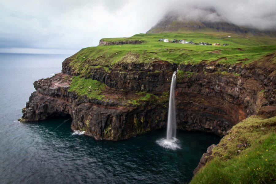 Faroe Islands coastal cliff waterfall with grassy plateau and small village.