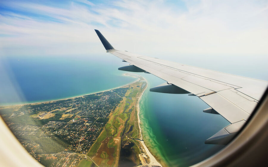Aerial view of airplane wing over peninsula, ocean, and town under a sunny sky.
