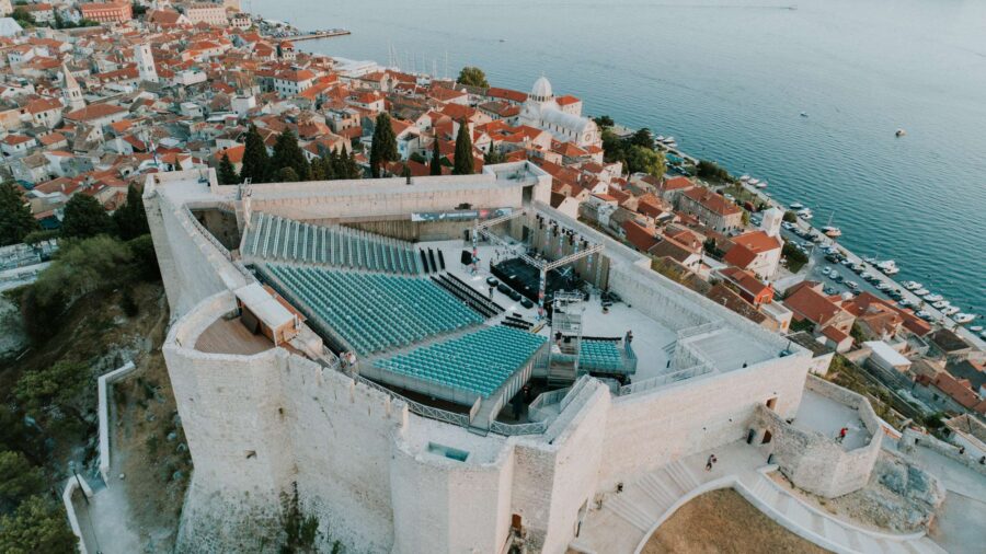 Aerial view of coastal amphitheater in ancient fortress with terracotta rooftops and harbor.