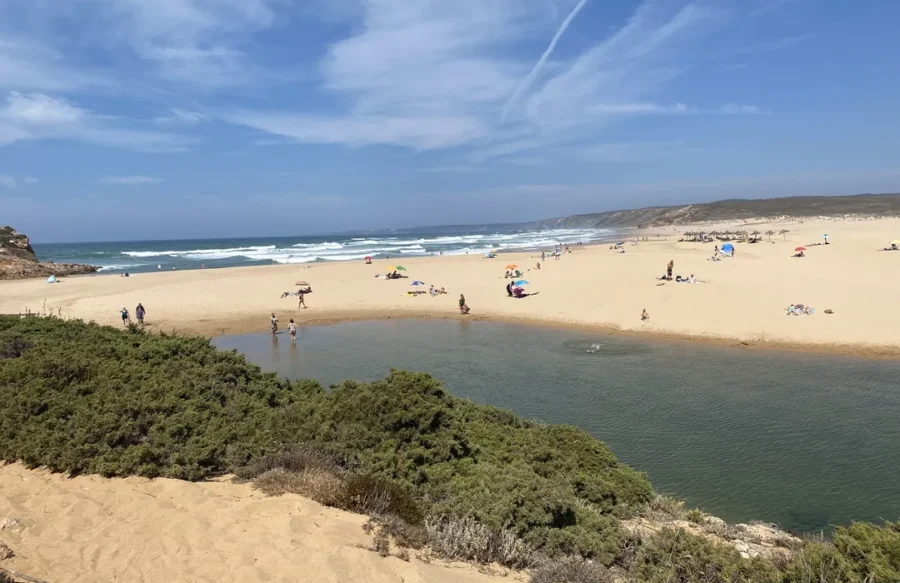 Pontal da Carrapateira beach: golden sand dunes, tidal lagoon, ocean waves, beachgoers, blue sky.