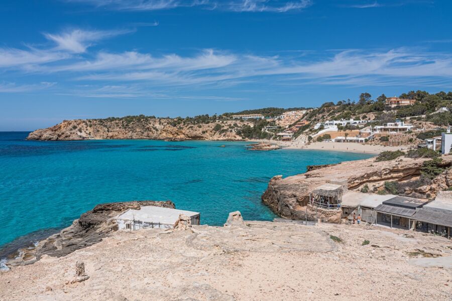 Coastal Cala Tarida beach scene with cliffs, sandy shore, blue sea, and beachfront café.