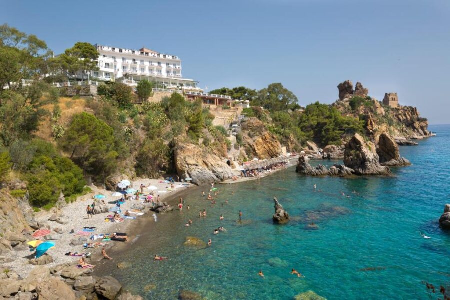 Mediterranean beach scene with sunbathers, rocky shoreline, and a hillside hotel.