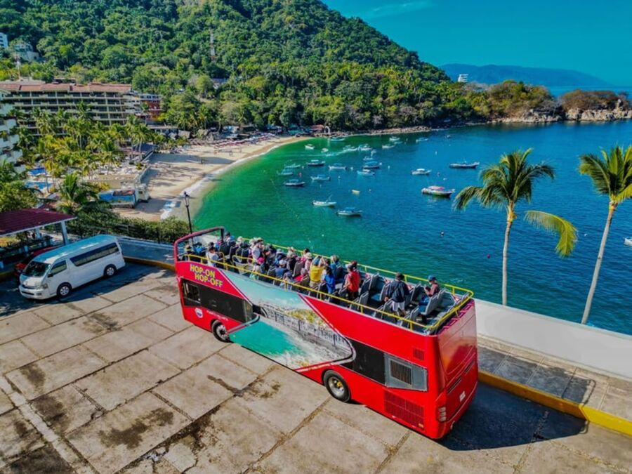 Coastal tour with double-decker bus, tourists, picturesque bay, and vibrant tropical scenery.