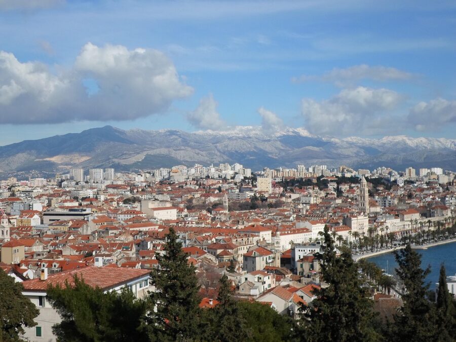 Vibrant coastal city skyline with mountains, terracotta rooftops, and blue harbor waters.