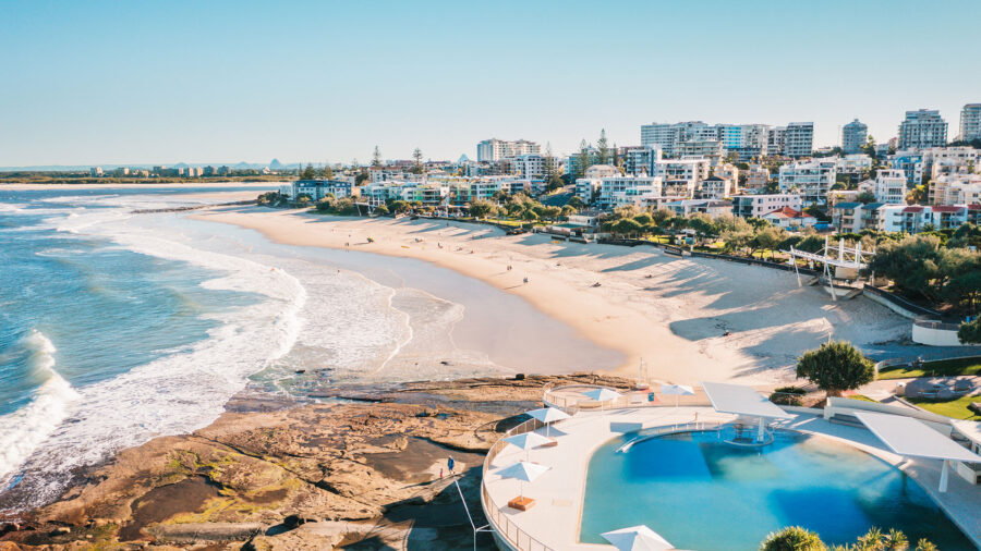 Aerial view of Caloundra beach, oceanfront pool, coastal city skyline and waves.