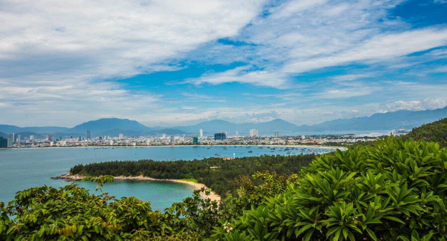 Panoramic view of Da Nang Bay with lush greenery, sandy beach, coastal city skyline and mountains.