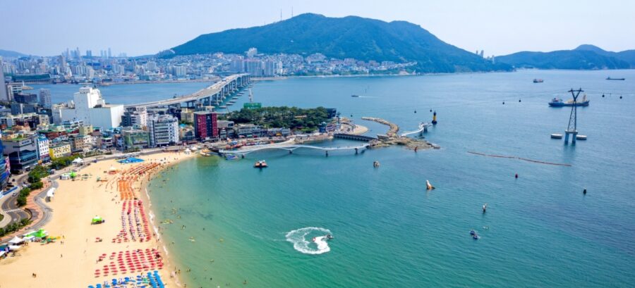 Aerial view of Songdo Beach Busan with curved sandy shoreline, umbrellas, city skyline and mountains.