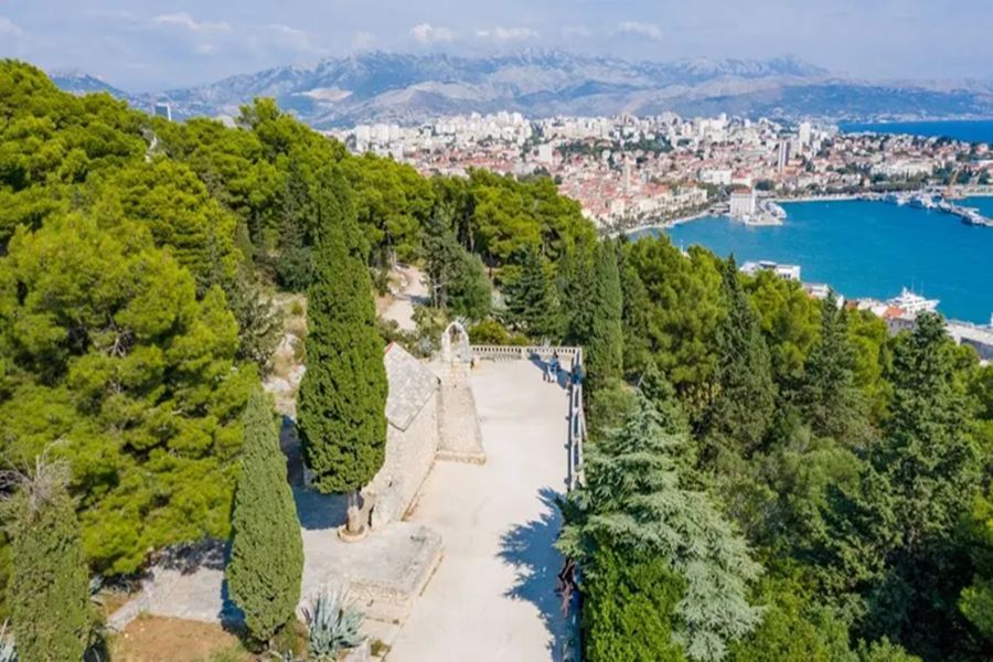 Aerial view of coastal city with lush hills, historic architecture, and sparkling blue sea.
