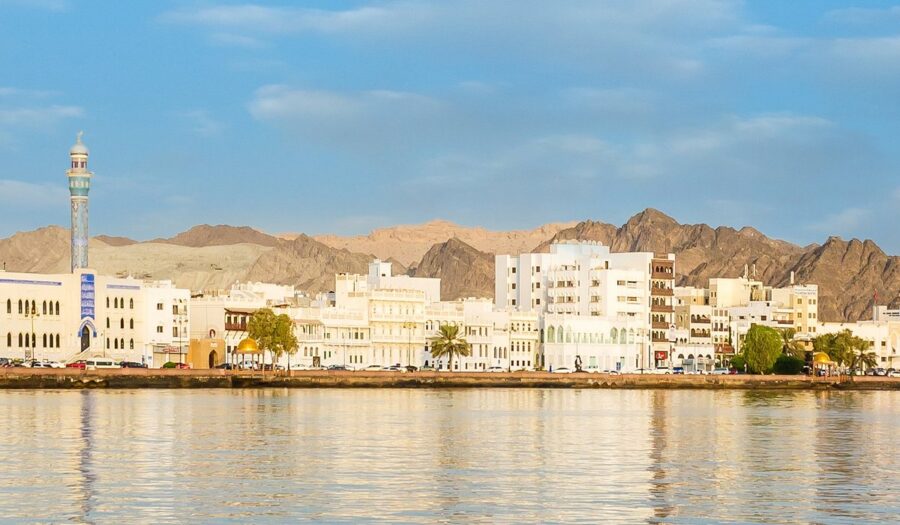Waterfront view of Muscat Oman with mosque minaret, white buildings and rugged brown mountains.