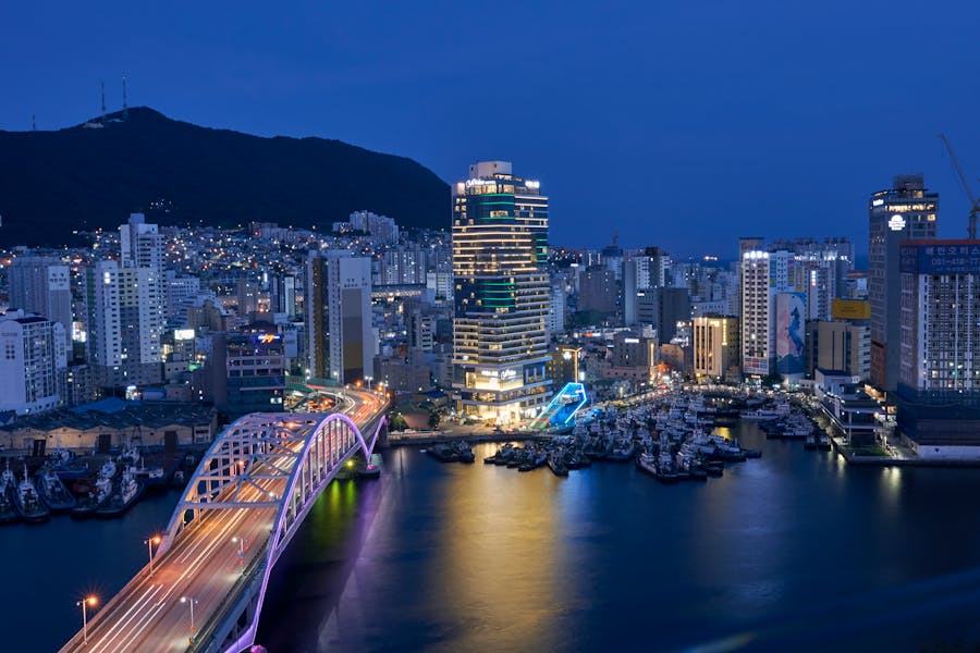 Vibrant coastal city harbor at dusk with illuminated skyscrapers, bridge, boats, and mountain backdrop.