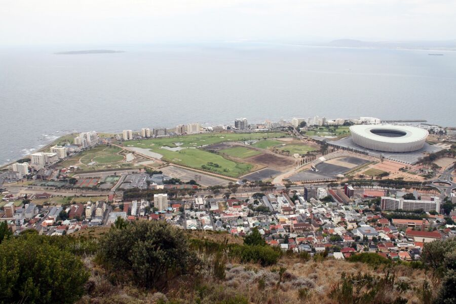 Aerial view of coastal city with vibrant buildings, stadium, green spaces, and ocean horizon.