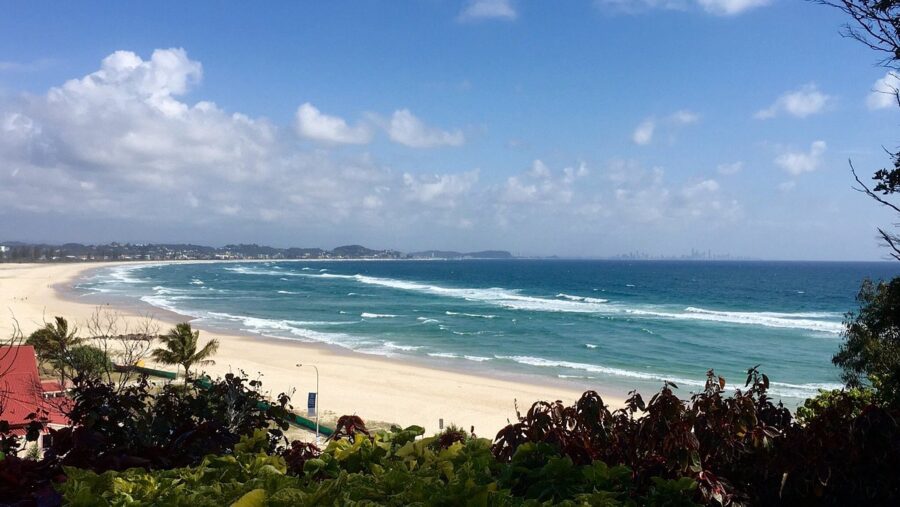 Panoramic Kirra Beach surf coastline with turquoise ocean, sandy shore, palm trees and distant city skyline.