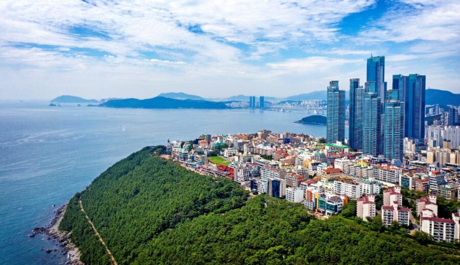 Coastal city skyline with green Dalmaji Hill, tall waterfront skyscrapers, ocean bay and distant bridge.