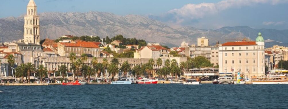 Picturesque coastal cityscape with historic tower, palm-lined promenade, cafes, and serene mountain backdrop.