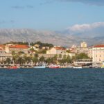 Picturesque coastal cityscape with historic tower, palm-lined promenade, cafes, and serene mountain backdrop.