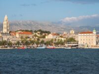 Picturesque coastal cityscape with historic tower, palm-lined promenade, cafes, and serene mountain backdrop.