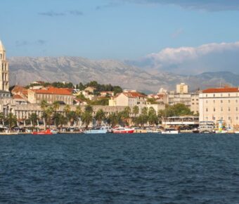 Picturesque coastal cityscape with historic tower, palm-lined promenade, cafes, and serene mountain backdrop.