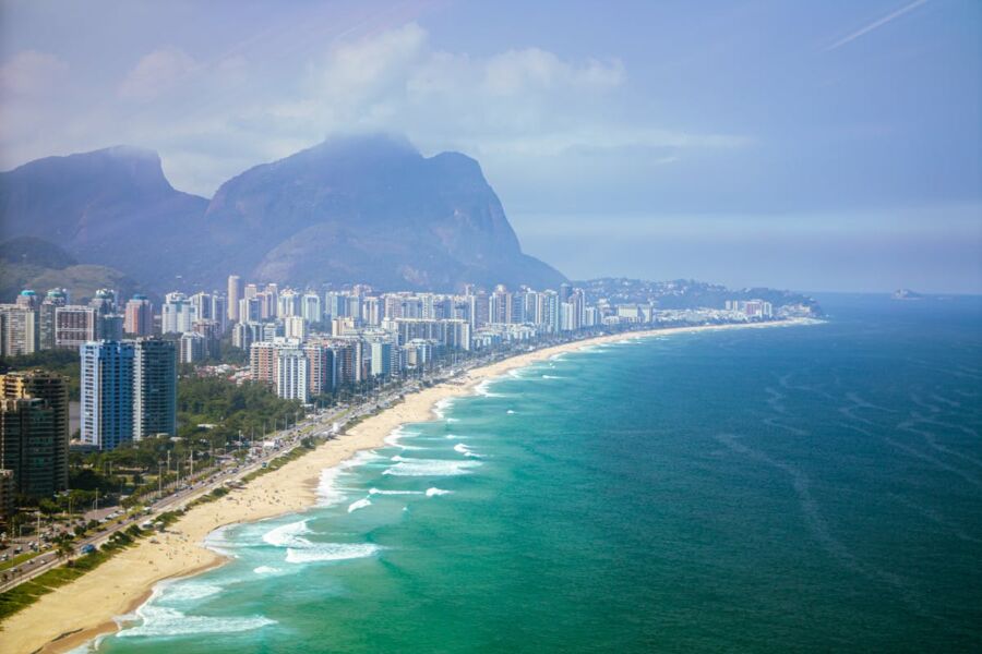Vibrant coastal cityscape with beach, high-rises, palm trees, and mountains under blue sky.
