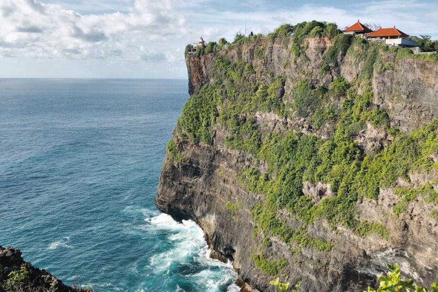Dramatic coastal Uluwatu cliff features tropical vegetation, Balinese buildings, and turquoise ocean.