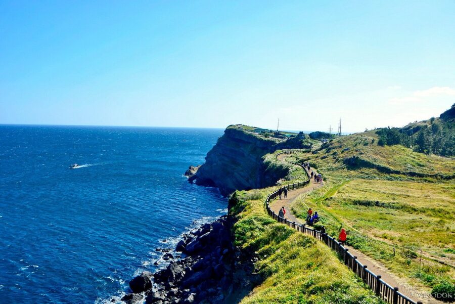 Scenic coastal cliff walking trail overlooking blue ocean, grassy headlands, hikers and rocky shoreline.