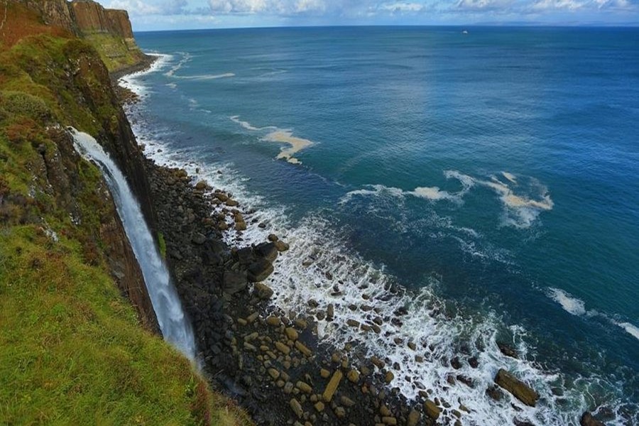 Waterfall cascading from lush cliffs into ocean, surrounded by rugged coastline and vibrant greenery.