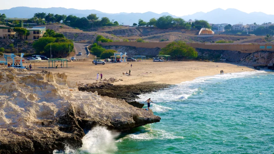 Scenic Qurum beach with rocky cliffs, turquoise sea, golden sand, and distant mountains.