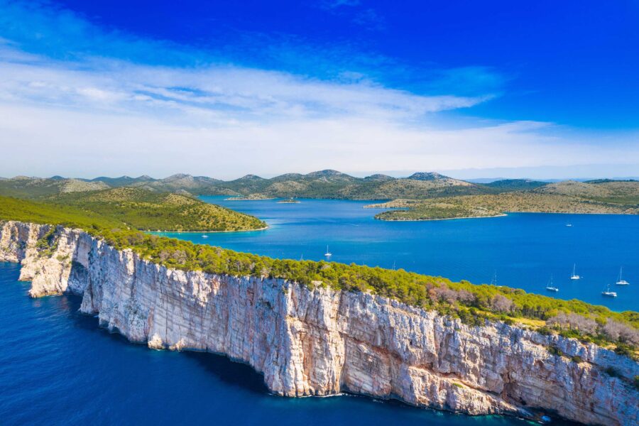 Rugged white cliffs and azure sea with lush greenery, boats, and islands under a bright sky.