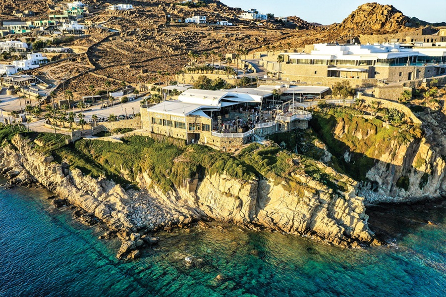 Aerial view of Greek island cliffside restaurant with turquoise sea and modern villas.