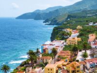 Colorful Mediterranean cliffside town with palm trees overlooking turquoise ocean and dramatic rocky coastline.