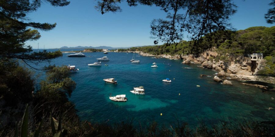 Cap d’Antibes coastal cove with turquoise water, moored boats, yachts, and pine-fringed rocky shore.