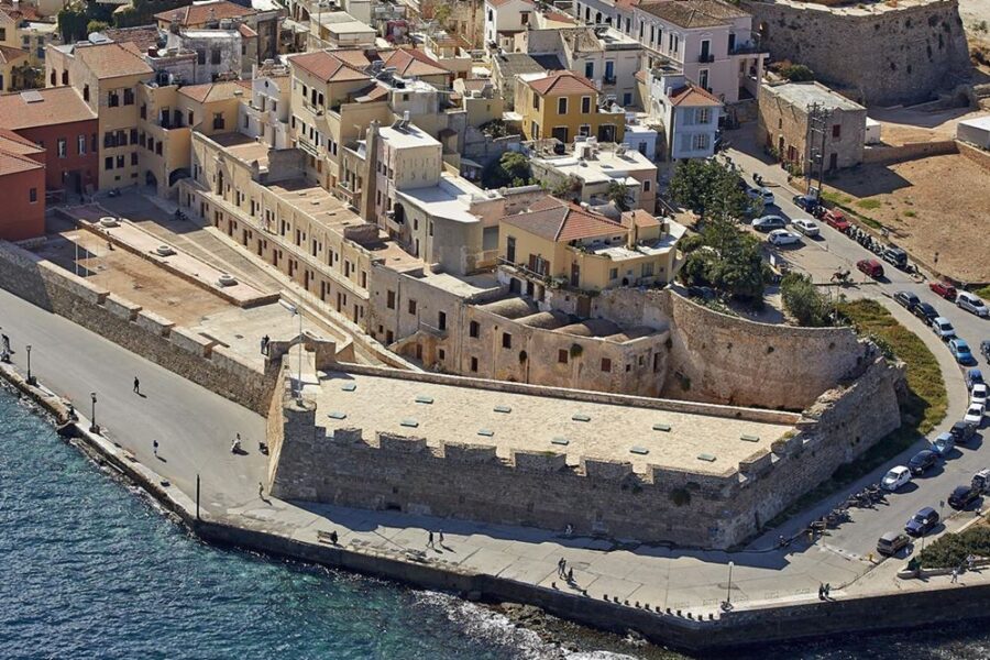 Historic coastal fortress and seaside promenade overlooking turquoise sea and terracotta-roofed Mediterranean town.