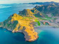 Aerial view of desert coastal golf resort peninsula with sandy beach, rocky cliffs and turquoise sea.