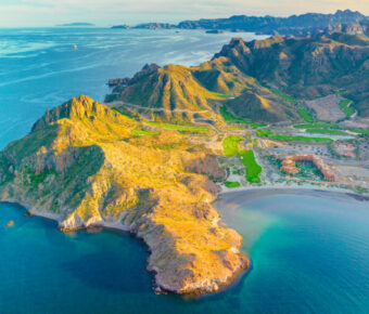 Aerial view of desert coastal golf resort peninsula with sandy beach, rocky cliffs and turquoise sea.