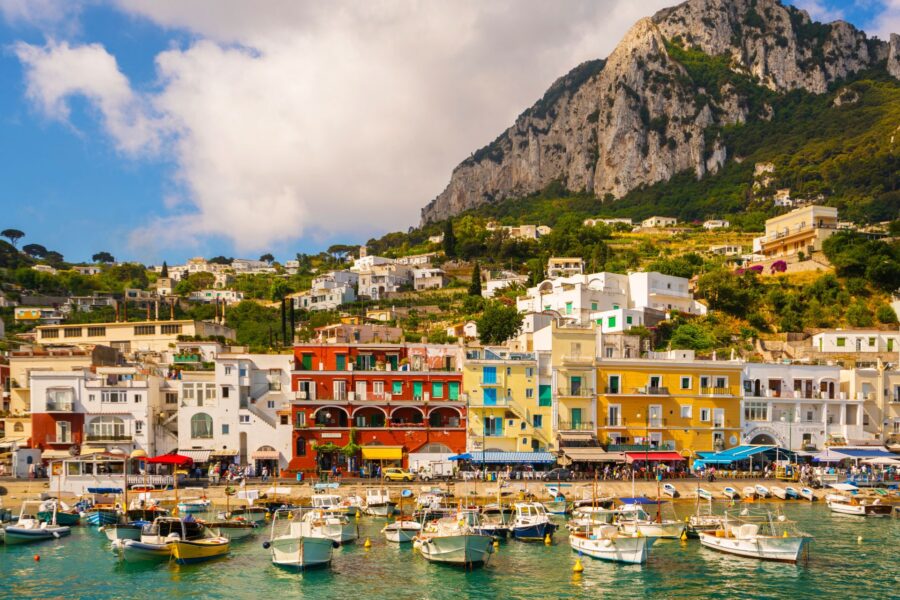 Colorful Mediterranean harbor with moored boats, waterfront cafes, and cliffside houses.