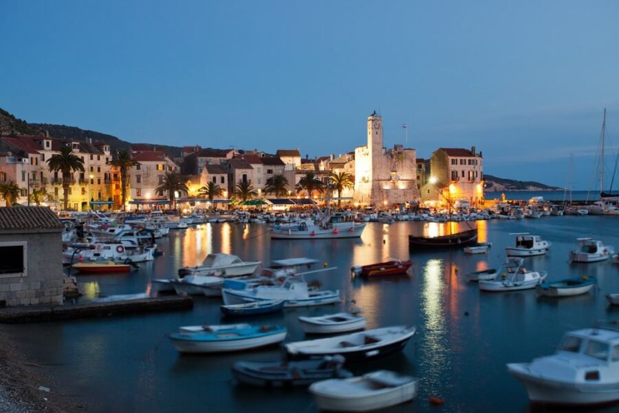Mediterranean harbor at dusk with moored boats, waterfront restaurants, illuminated stone tower.