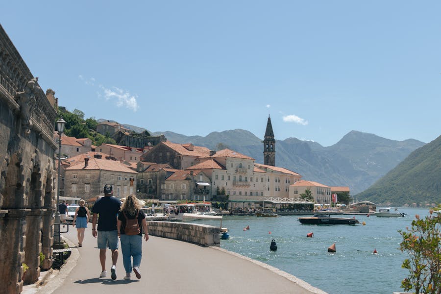 Kotor, Montenegro harbor promenade with couple walking, historic stone buildings and moored boats.