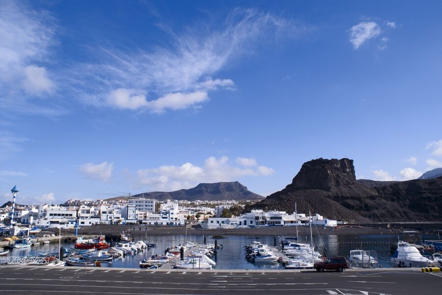 Scenic coastal harbor with boats, white buildings, blue sky, and dramatic mountains.