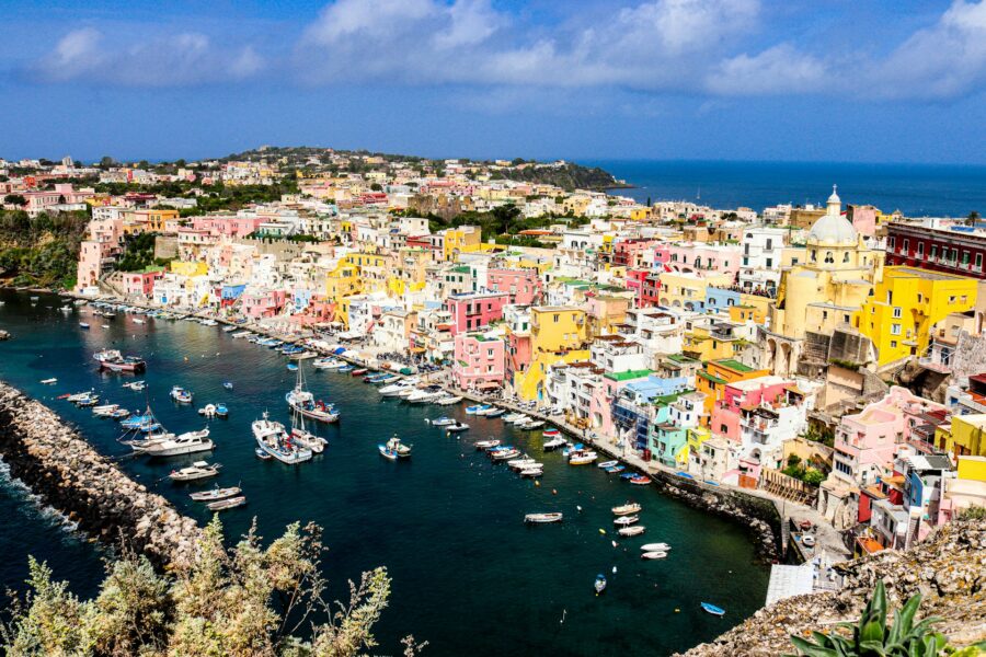 Mediterranean coastal harbor town with colorful buildings, boats, and stone breakwater.
