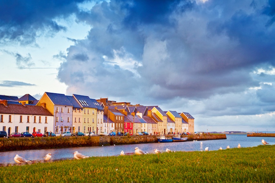 Colorful coastal houses on Galway quay, Ireland, with tidal channel, seagulls, dramatic sky.