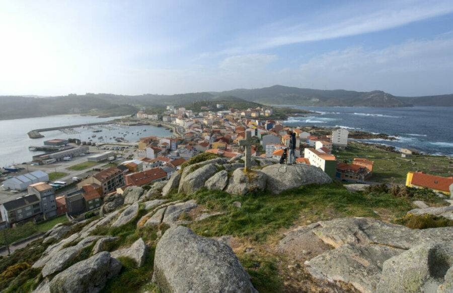 Coastal harbor village seen from rocky viewpoint with photographer, red-tiled roofs and ocean bay.