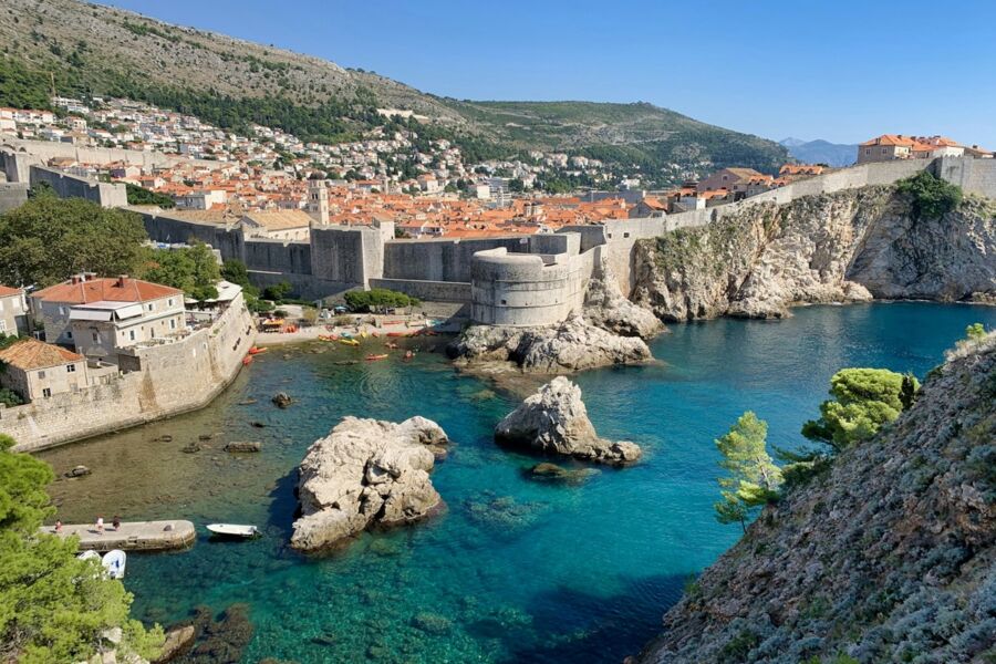 Coastal Dubrovnik historic town with fortified walls, orange rooftops, turquoise waters, and lush green hills.