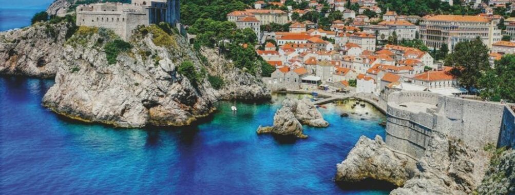 Scenic Croatia coastal landscape with historic castle, rocky shore, vibrant village, blue sea, and rolling hills.