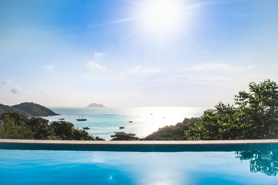 Luxury infinity pool overlooking Búzios coastline, turquoise ocean, sailboats and lush tropical hills at sunset.