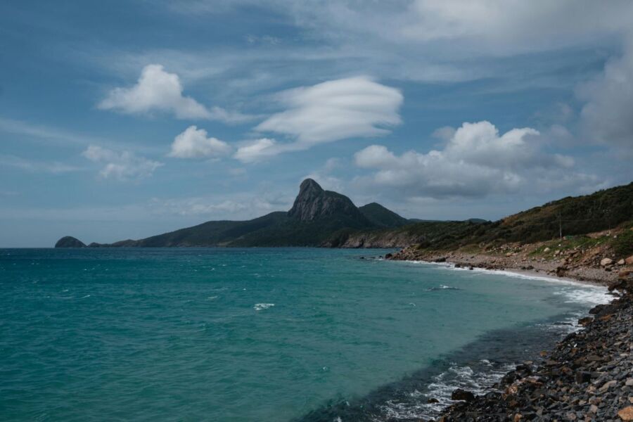 Turquoise coastal scene with mountain, pebbled beach, and dynamic sky.