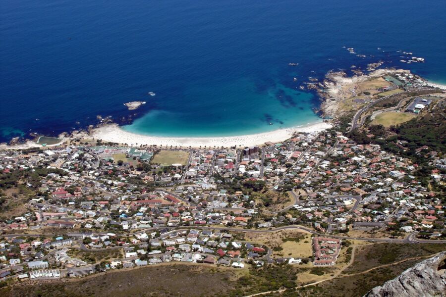 Aerial view of coastal landscape with sandy beach, urban development, and scenic mountains.