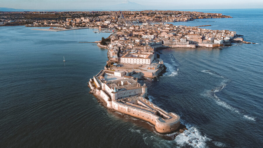 Aerial view of Ortygia, showcasing Mediterranean architecture, fortress, and Mount Etna backdrop.