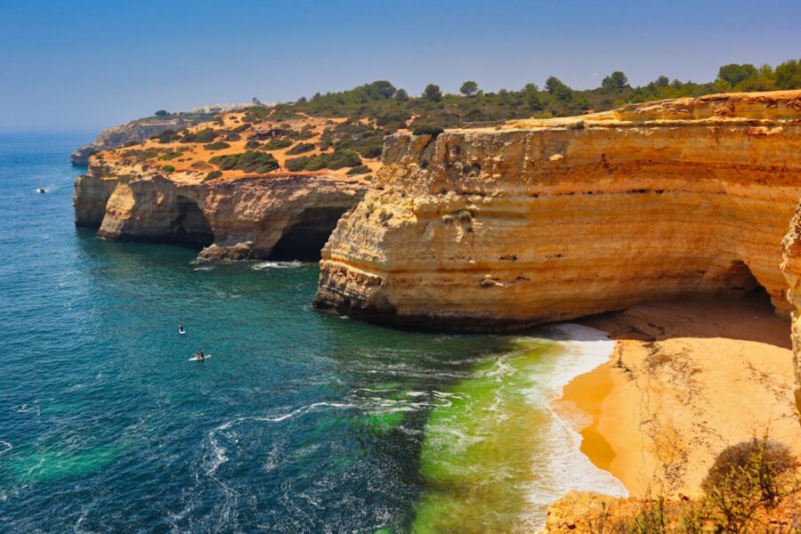 Dramatic Lagos coastal cliffs with blue waters, sandy beach, boats, and lush vegetation.