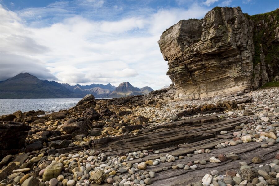 Rugged coastal scene with pebbles, cliffs, and distant mountains under a clear blue sky.